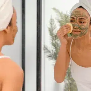 A woman with a towel-wrapped head applies a clay face mask while looking in a mirror. She gently uses a sponge, conveying a calm self-care routine.