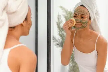 A woman with a towel-wrapped head applies a clay face mask while looking in a mirror. She gently uses a sponge, conveying a calm self-care routine.