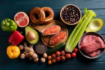 A variety of fresh foods: salmon, steak, colorful bell peppers, tomatoes, celery, grapefruit, avocados, beans, walnuts, bagels, cucumber, chia seeds, and ginger against a dark blue wooden background.