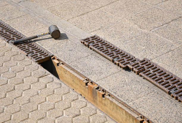 A hammer rests beside a partially opened storm drain with metal grating on a paved sidewalk. The tone suggests maintenance or repair work.