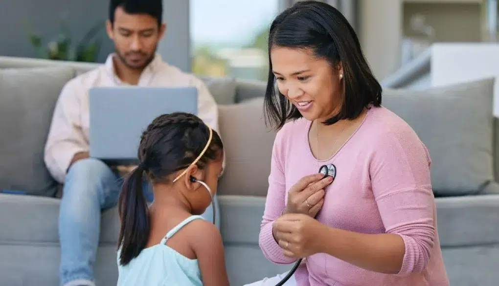 Woman in pink uses a stethoscope to listen to a young girl’s chest while a man works on a laptop in the background.