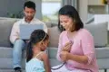 Woman in pink uses a stethoscope to listen to a young girl’s chest while a man works on a laptop in the background.