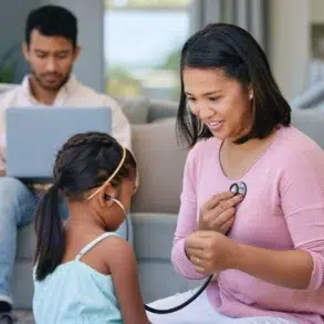 Woman in pink uses a stethoscope to listen to a young girl’s chest while a man works on a laptop in the background.