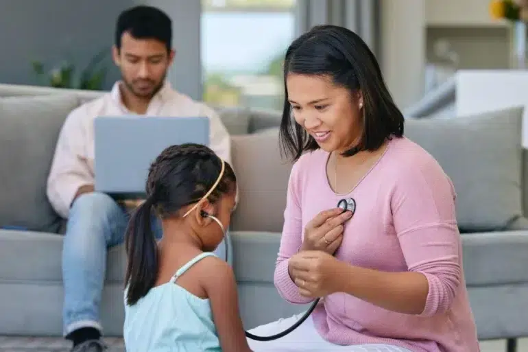 Woman in pink uses a stethoscope to listen to a young girl’s chest while a man works on a laptop in the background.