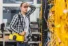 A man in a checkered shirt stands in a server room holding a large yellow device, looking thoughtful. Tangled yellow cables fill the right side.