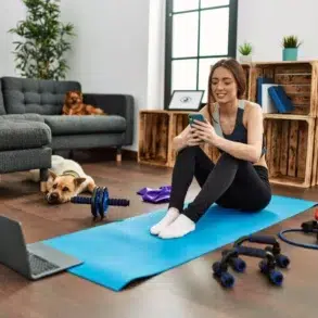 Woman sitting on a blue yoga mat in a living room, smiling at her phone. Exercise equipment and a laptop are nearby. A dog rests on the floor. Cozy and relaxed atmosphere.