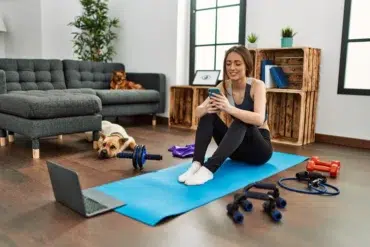 Woman sitting on a blue yoga mat in a living room, smiling at her phone. Exercise equipment and a laptop are nearby. A dog rests on the floor. Cozy and relaxed atmosphere.