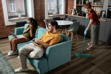 Two people sit on a teal sofa; one uses a laptop, the other plays video games. In the background, another person mops the modern kitchen floor. Casual and relaxed setting.