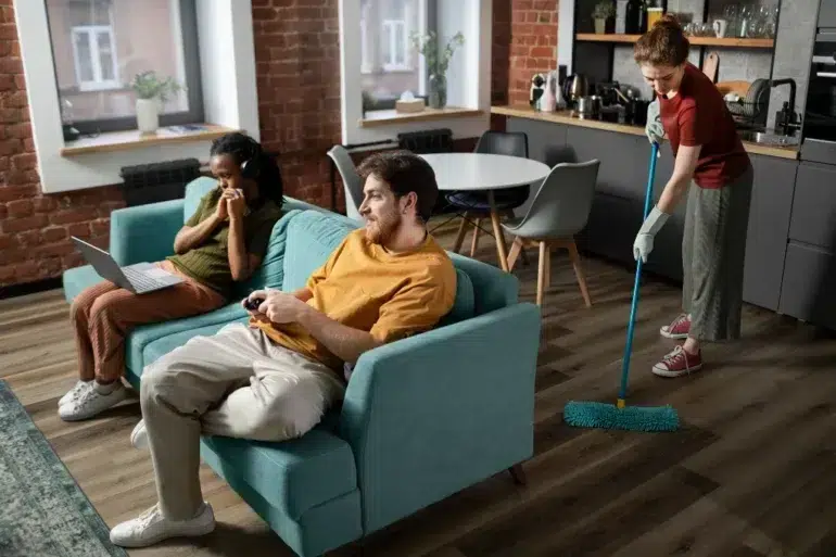 Two people sit on a teal sofa; one uses a laptop, the other plays video games. In the background, another person mops the modern kitchen floor. Casual and relaxed setting.