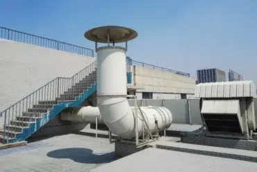 Rooftop scene with industrial ventilation pipes and exhaust systems against a clear blue sky. A staircase with a blue railing is on the left.