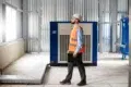 A man wearing a hard hat and orange safety vest inspects industrial equipment in a modern facility. He holds a black folder, appearing focused.