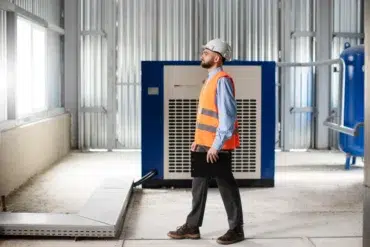 A man wearing a hard hat and orange safety vest inspects industrial equipment in a modern facility. He holds a black folder, appearing focused.