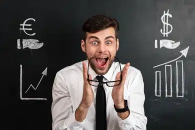 A man in a white shirt and tie looks excited, holding glasses. Behind him is a blackboard with chalk drawings of currency symbols and rising graphs.