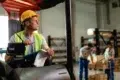 Man in a forklift wearing a yellow hard hat and safety vest, holding a pen and clipboard, looks focused in a warehouse. Workers in background handle boxes.
