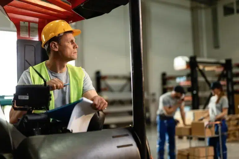 Man in a forklift wearing a yellow hard hat and safety vest, holding a pen and clipboard, looks focused in a warehouse. Workers in background handle boxes.