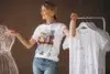 Smiling woman wearing a printed white T-shirt and jeans. She stands between a wire mannequin and a rack of minimalist T-shirts, with a relaxed vibe.