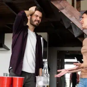 A man and woman engage in a lively conversation in a kitchen. The man looks thoughtful and the woman gestures expressively. Red cups are on the counter.