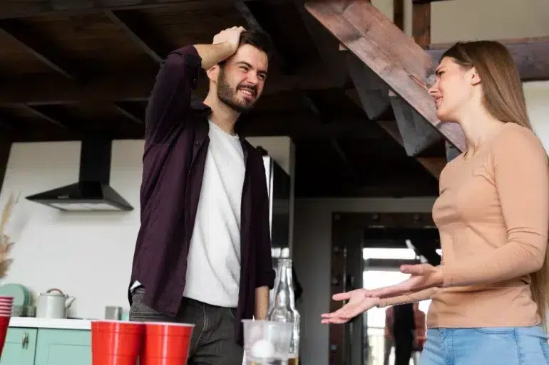 A man and woman engage in a lively conversation in a kitchen. The man looks thoughtful and the woman gestures expressively. Red cups are on the counter.