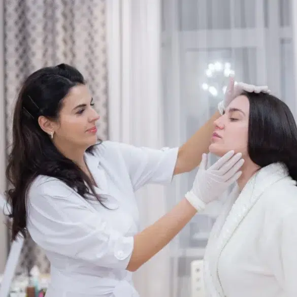 A cosmetologist in a white coat examines a woman's face in a bright, softly-lit room. The woman wears a white robe. The scene feels calm and professional.