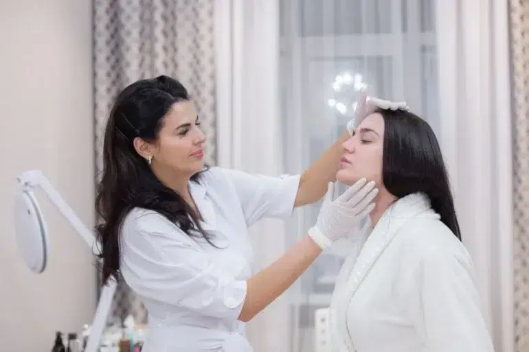 A cosmetologist in a white coat examines a woman's face in a bright, softly-lit room. The woman wears a white robe. The scene feels calm and professional.