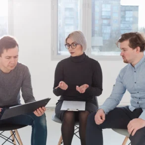 Three people sit in a bright room, engaged in discussion. The woman in the center gestures with her hands, while the men listen intently.