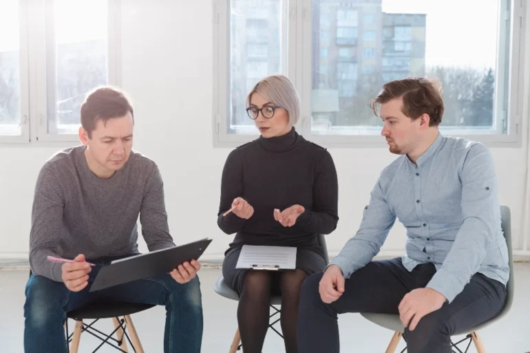 Three people sit in a bright room, engaged in discussion. The woman in the center gestures with her hands, while the men listen intently.