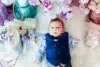 A baby in a blue vest lies on a white fluffy rug, surrounded by pastel holiday gifts and a white Christmas tree, creating a festive, joyful atmosphere.