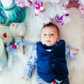 A baby in a blue vest lies on a white fluffy rug, surrounded by pastel holiday gifts and a white Christmas tree, creating a festive, joyful atmosphere.