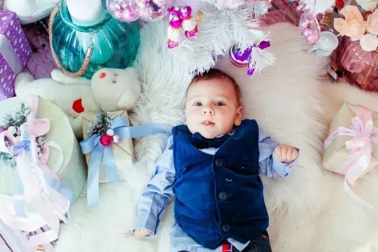 A baby in a blue vest lies on a white fluffy rug, surrounded by pastel holiday gifts and a white Christmas tree, creating a festive, joyful atmosphere.