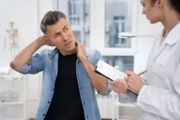 A man looking pained touches his neck while discussing with a female doctor holding a clipboard in a bright medical office.