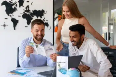 Three people in an office, smiling and looking at travel brochures. A world map is on the wall, and a small globe sits on the table, conveying excitement.
