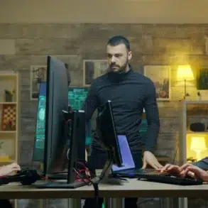 Three people collaborate in an office with computers and screens. A serious atmosphere prevails as they focus on their tasks at a shared desk.