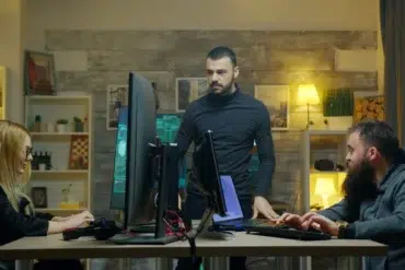 Three people collaborate in an office with computers and screens. A serious atmosphere prevails as they focus on their tasks at a shared desk.