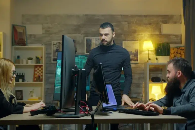Three people collaborate in an office with computers and screens. A serious atmosphere prevails as they focus on their tasks at a shared desk.