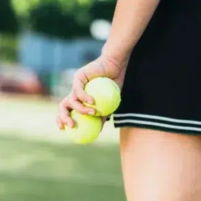 A person in a black skirt, holding two yellow tennis balls near their side on a sunlit court, suggesting readiness and focus.