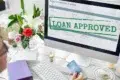 Computer screen showing an application form with "LOAN APPROVED" in bold green letters. Nearby, a person holds a credit card, with flowers on the desk.