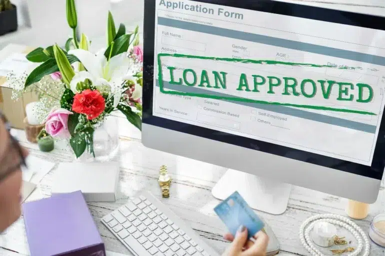 Computer screen showing an application form with "LOAN APPROVED" in bold green letters. Nearby, a person holds a credit card, with flowers on the desk.