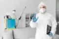 Two people in full protective suits and gloves disinfecting a living room, one holding a clipboard and raising a hand.