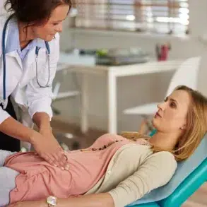 A doctor performs an abdominal examination on a patient lying on a medical table in a bright, airy clinic. The atmosphere is professional and caring.