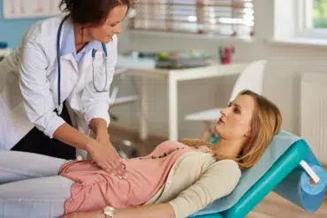 A doctor performs an abdominal examination on a patient lying on a medical table in a bright, airy clinic. The atmosphere is professional and caring.
