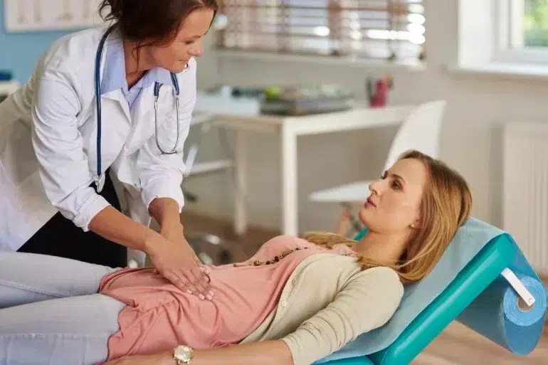 A doctor performs an abdominal examination on a patient lying on a medical table in a bright, airy clinic. The atmosphere is professional and caring.