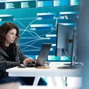 A woman works intently on a laptop at a desk, with a digital network display on the wall behind her. The setting is modern and technological.