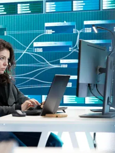 A woman works intently on a laptop at a desk, with a digital network display on the wall behind her. The setting is modern and technological.