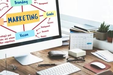 Computer screen displaying a marketing diagram with branches labeled "Branding," "Goals," "Vision." Desk with calendar, keyboard, and plant nearby.