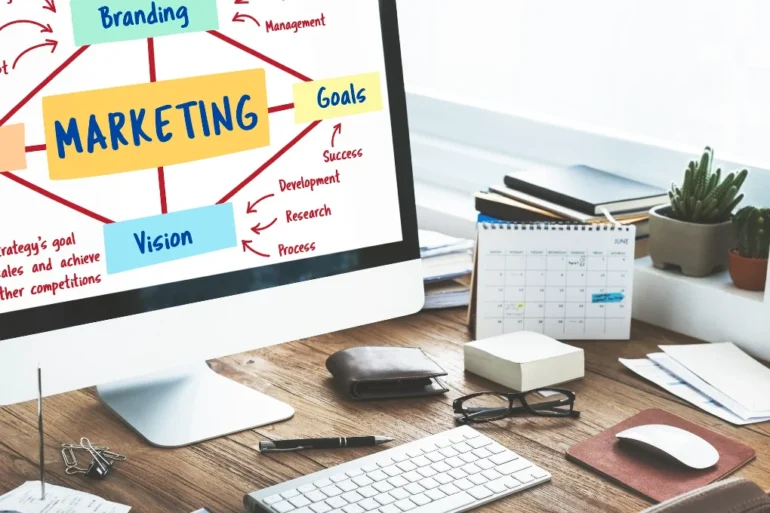 Computer screen displaying a marketing diagram with branches labeled "Branding," "Goals," "Vision." Desk with calendar, keyboard, and plant nearby.