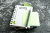 Stack of notebooks on a textured dark surface with a pen, blank sticky notes, and green push pins on top.