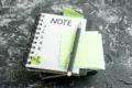 Stack of notebooks on a textured dark surface with a pen, blank sticky notes, and green push pins on top.
