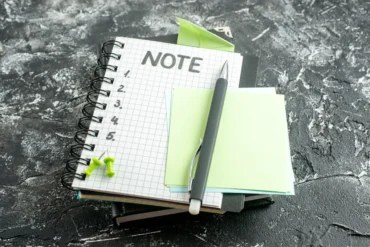 Stack of notebooks on a textured dark surface with a pen, blank sticky notes, and green push pins on top.
