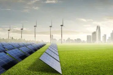 A field of solar panels and wind turbines stand on a grassy plain, with a city skyline in the background under a bright, partly cloudy sky.