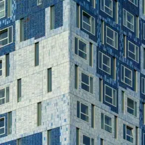 Close-up view of a modern building with mosaic tiles in varying shades of blue and white. The geometric pattern creates a textured, dynamic facade with many windows.